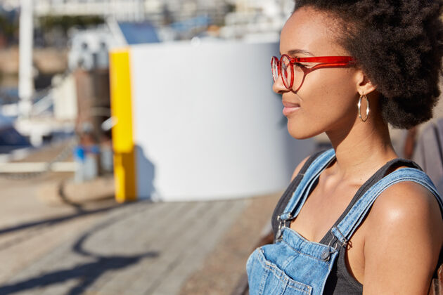 眼镜非洲式发型 戴眼镜 牛仔工作服的快乐黑人女青年侧身户外镜头 聚焦在远方 暑假曾在户外散步 漫步在未知的城市街道肖像非洲