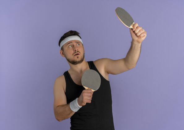 健康戴着头巾拿着两个球拍打乒乓球的年轻健身男子站在紫色的墙上困惑地看着一边教练靠边站男