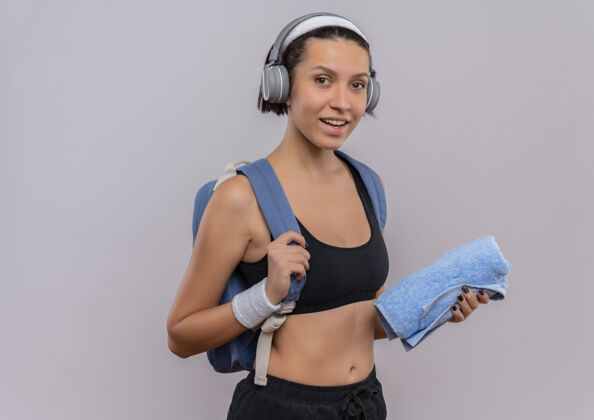 站着穿着运动服的年轻健身女士 背着背包 头上戴着耳机 手拿毛巾 站在白色的墙壁上 微笑着积极快乐健康姿势年轻