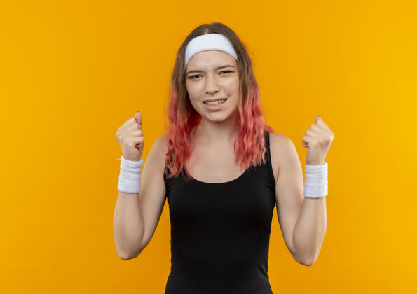 女性身着运动服的年轻健身女士握紧拳头 站在橙色的墙上 高兴地欢呼着自己的成功高兴人快乐