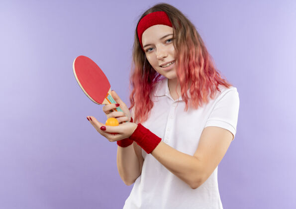 年轻人戴着头巾 拿着乒乓球球拍 脸上带着微笑的年轻女子站在紫色的墙上运动装球抱