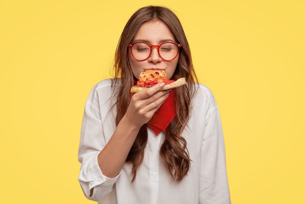 味道嗯 那么美味的黑发美女吃意大利披萨片 闭上眼睛不高兴 享受美味 戴着眼镜和衬衫 隔离在黄色的墙壁上吃的概念披萨店热的快餐