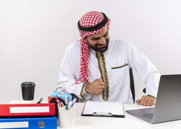 拳头身着传统服装的阿拉伯商人坐在桌旁 手提电脑握紧拳头 高兴地离开了办公室工作电脑商务人士