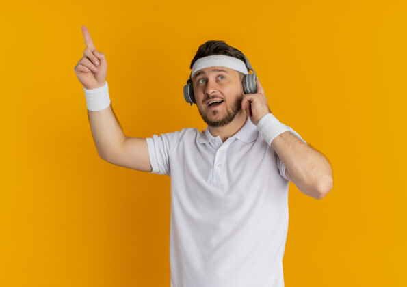 健身身穿白衬衫 头戴耳机的年轻健身男子站在橘红色的墙上 惊讶而高兴地伸出食指 有着伟大的想法运动耳机运动