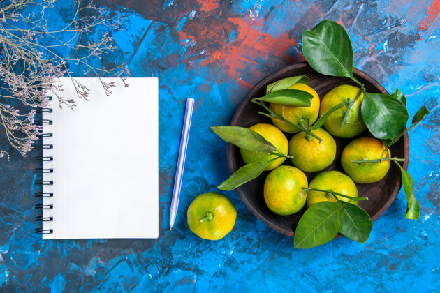 笔记本顶视图黄色的橘子叶子放在木制的碗里一个笔记本和紫色的钢笔放在蓝色的表面上水果钢笔顶部