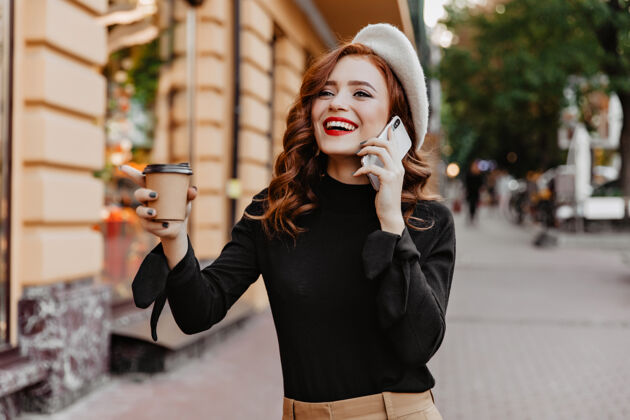 街头兴奋的红发女孩在街上喝咖啡迷人的时髦女人在城墙上打电话微笑成人肖像
