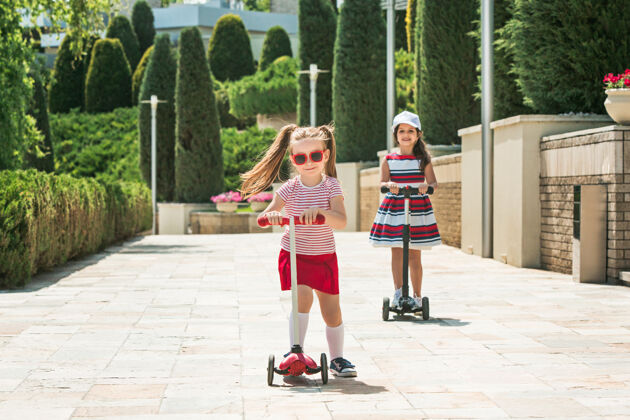 童年阳光明媚的日子里的女朋友们学龄前女孩们在户外骑着滑板车平衡街道户外