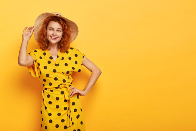 享受快乐夏日女士一只手放在腰上 另一只手戴在草帽上 穿着鲜艳的黄色圆点连衣裙 有着快乐的外表 苗条的身材 室内模特 为您的推广复制空间美丽和女人味时尚能量人