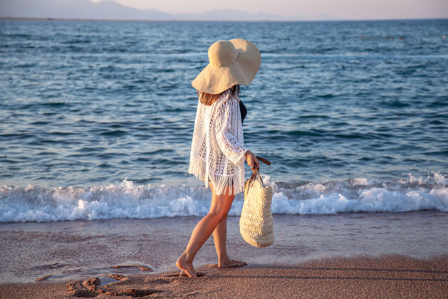 时尚一个戴着大帽子和柳条袋的女孩沿着海边散步暑假概念海滩波西米亚度假