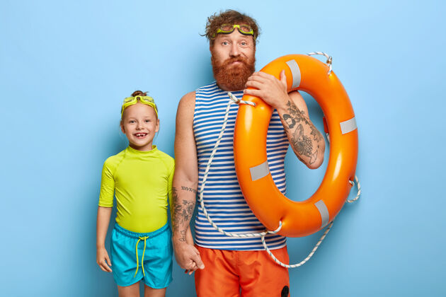 微笑家庭娱乐胡子姜爸爸牵着小女儿的手 穿着夏装 拿着游泳器材 在海边度暑假 孤零零的在蓝色的墙上 像这个季节团结女孩爸爸