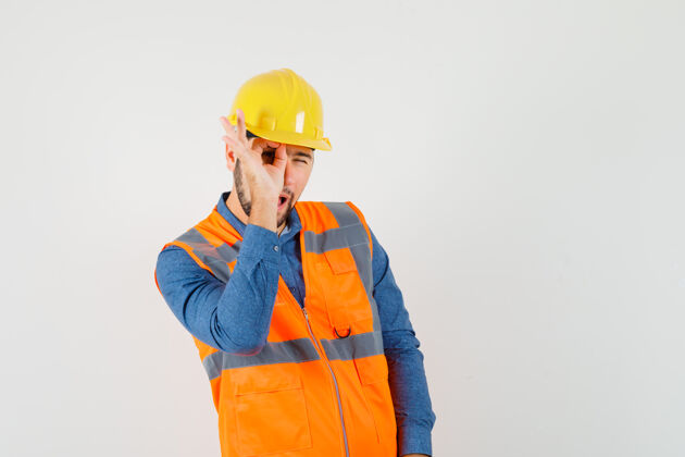 工具年轻的建筑工人穿着衬衫 背心 戴着头盔 眼睛上有“ok”的标志 看上去很好奇 正前方的景色工业安全安全帽