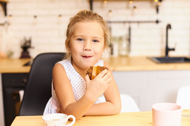 幸福可爱快乐的小女孩坐在舒适的厨房里吃着美味的饼干 餐桌上放着杯子可爱有趣的高加索宝宝愉快地嚼着烤甜饼饥饿糕点美味