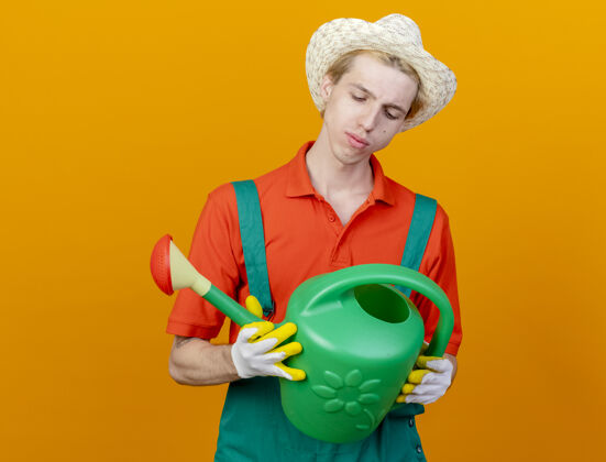 男人年轻的园丁穿着连体衣戴着帽子拿着喷壶站在橙色的背景下严肃地看着它穿着浇水看