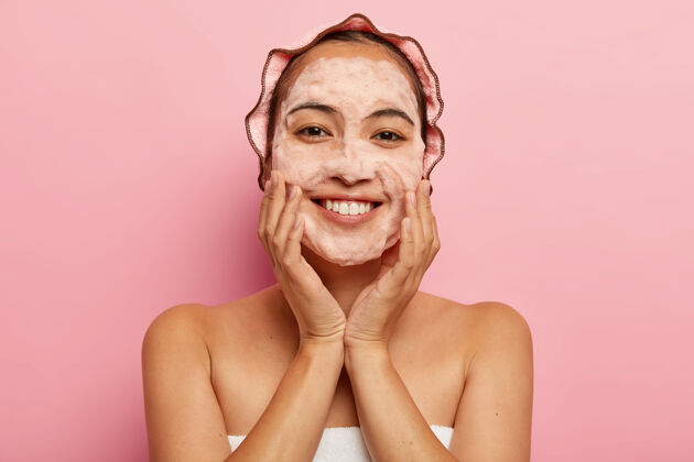 亚洲人韩国年轻女性的头像接触到无暇柔软的皮肤 用带有泡沫清洁剂的卫生肥皂洗脸 用毛巾包裹 头上有浴帽 隔离在粉色墙壁上清洁理念护理室内玫瑰色