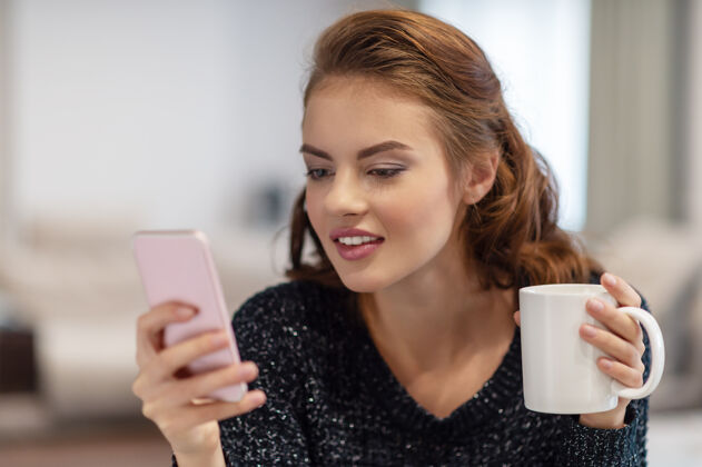 微笑漂亮的年轻女子拿着一杯咖啡看着她的智能手机女子在智能手机上输入信息迷人房间杯子
