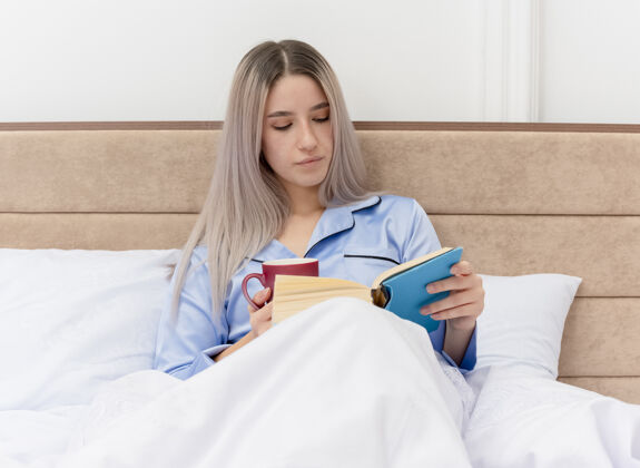 床穿着蓝色睡衣的年轻漂亮女人坐在床上 在卧室的室内灯光背景下 拿着一杯咖啡看书坐着蓝色咖啡