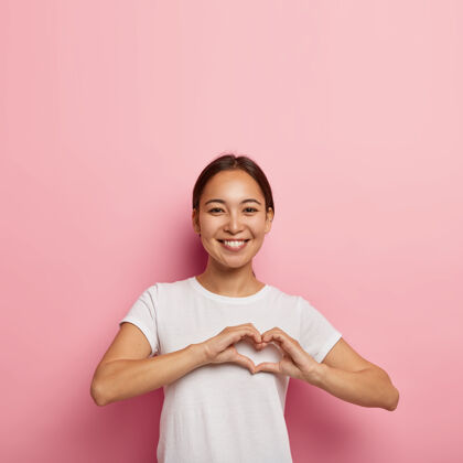 浪漫迷人的亚洲女性做心形手势 表达爱意 说是我的情人 积极微笑 穿白色服装 以空旷的空间对着粉色墙壁摆姿势肢体语言概念罗西自由空间心