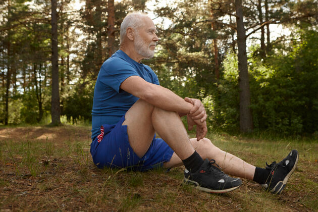 人时尚的男性退休者侧视图 留着胡须 坐在森林边欣赏美丽的风景 在早晨的有氧运动后放松 双臂抱住膝盖 拥有平静的神情放松放松成年人