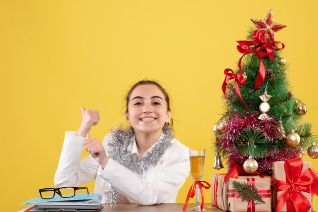 圣诞节前视图：女医生坐在桌子后面 拿着圣诞礼物和黄色背景上的树女快乐背景