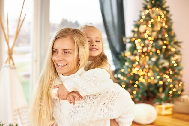 女人室内拍摄的快乐的年轻女子与长发给她可爱的小女儿骑小猪回来 玩得开心 在客厅里玩装饰闪闪发光的圣诞树骑车快乐孩子