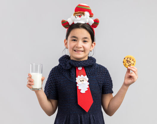 饼干穿着针织连衣裙的快乐小女孩戴着红色领带 头上戴着有趣的圣诞戒指 手里拿着一杯牛奶和饼干 开心地笑着连衣裙圣诞快乐新