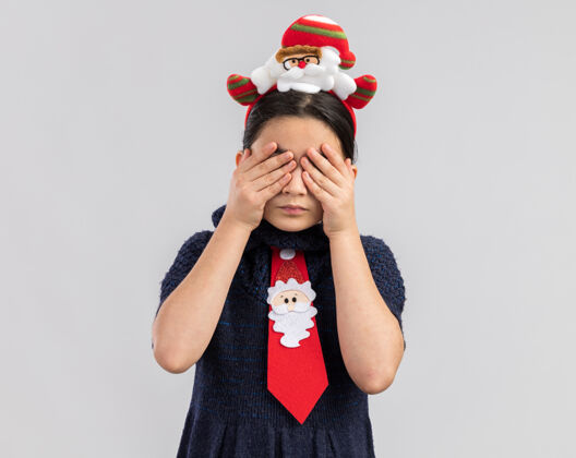 新小女孩穿着针织连衣裙 头上系着一条红色领带 上面戴着一个有趣的圣诞戒指 手捂着眼睛领带快乐手