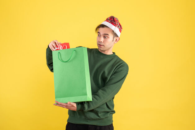 抱着正面图：戴着圣诞帽的年轻人拿着绿色的购物袋和礼物站在黄色的地板上好奇男人帽子