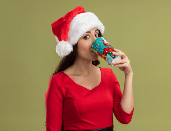 漂亮印象深刻的年轻漂亮女孩戴着圣诞帽拿着圣诞咖啡杯看着相机喝咖啡隔离在橄榄绿的背景女孩圣诞老人帽子