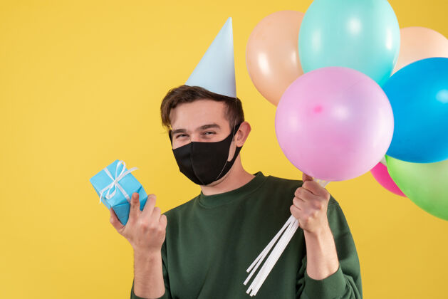 男人正面图：戴着派对帽和黑色面具的年轻人手里拿着蓝色礼品盒和黄色气球礼品盒表演者气球