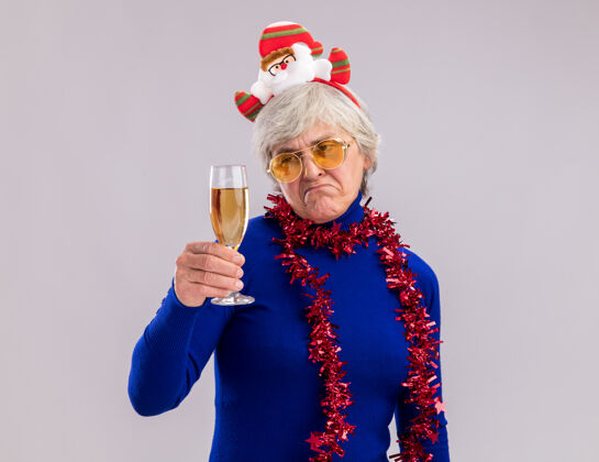 花环戴着太阳眼镜 脖子上戴着圣诞老人头带和花环的悲伤的老妇人抱着并看着一杯香槟酒 这杯香槟酒孤立地放在白色背景上 留着复印空间快乐悲伤一年
