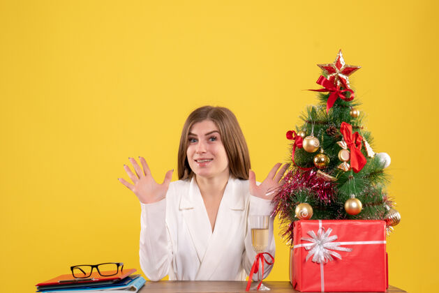 快乐前视图女医生坐在桌子前 带着圣诞礼物和黄色背景的树树圣诞节背景