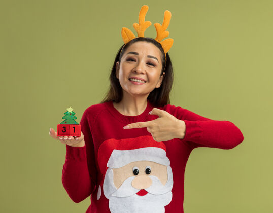 立方体穿着红色圣诞毛衣的快乐年轻女子 戴着有趣的鹿角边 展示着新年日期的玩具立方体 她愉快地笑着 用食指指着站在绿色背景上的立方体站立日期毛衣