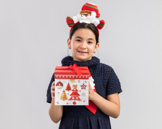 站着小女孩穿着针织连衣裙 头上系着红色领带 头上戴着有趣的圣诞戒指 手里拿着圣诞礼物 脸上带着微笑 快乐而积极穿边快乐