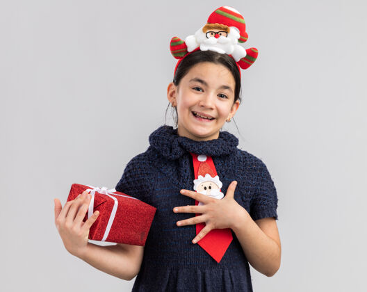 头穿着针织连衣裙的小女孩头上系着一条红色领带 头上戴着一个有趣的圣诞戒指 手里拿着圣诞礼物 面带微笑 心怀感激新的圣诞快乐微笑