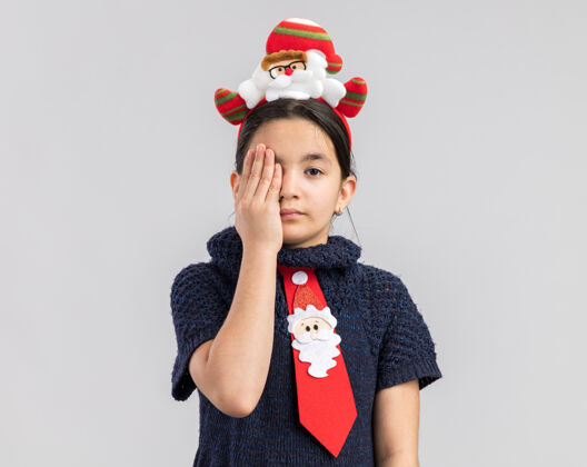 年小女孩穿着针织连衣裙 头上系着一条红色领带 戴着滑稽的圣诞戒指 一只手捂着一只眼睛 表情严肃脸穿一个