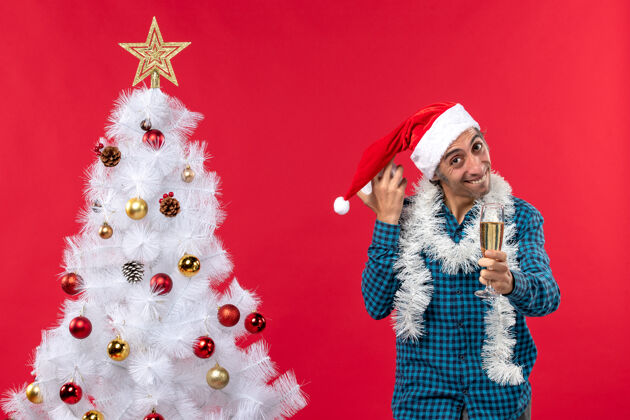 玻璃微笑快乐的年轻人玩着圣诞老人的帽子 举起一杯葡萄酒 拿着红色圣诞树旁的时钟庆祝玩圣诞