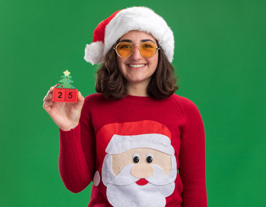 五穿着圣诞毛衣 戴着圣诞帽 戴着圣诞眼镜 手里拿着25号玩具立方体 脸上带着微笑 站在绿色的墙上戴着圣诞节圣诞老人