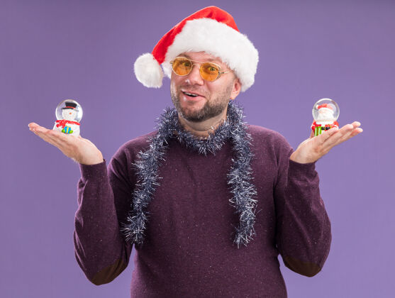 周围令人印象深刻的中年男子戴着圣诞帽 脖子上戴着金箔花环 戴着眼镜 把雪人和圣诞老人雕像孤立在紫色的墙上小雕像花环金属丝
