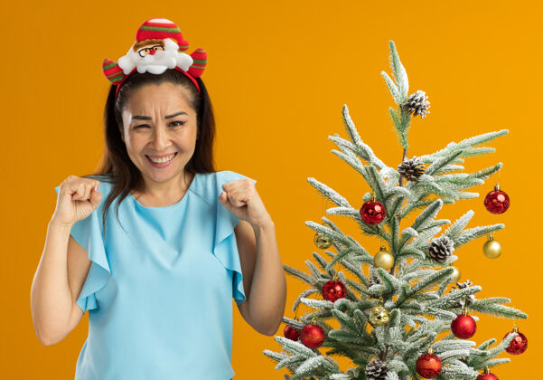 拳头身穿蓝色上衣的年轻女子头戴有趣的圣诞戒指 握紧拳头 站在橙色背景下的圣诞树旁 既高兴又兴奋头橙色兴奋
