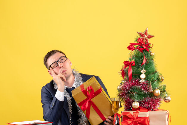 西装前视图困惑的人戴着眼镜坐在圣诞树附近的桌子上 黄色的礼物男性困惑圣诞节