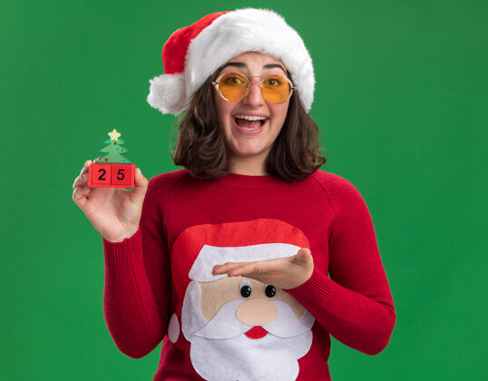 毛衣穿着圣诞毛衣的小女孩戴着圣诞帽戴着眼镜拿着25号玩具立方体微笑着高高兴兴地伸出手臂站在绿色的墙上穿着帽子圣诞老人