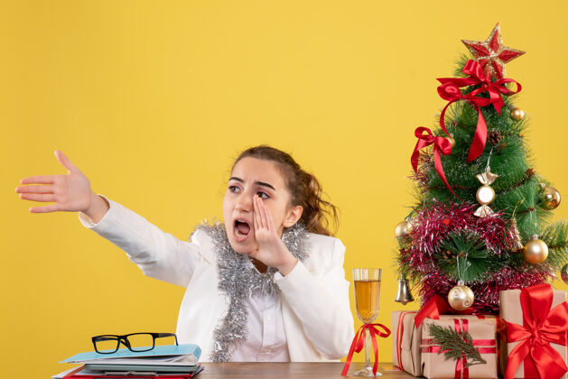 背景前视图：女医生坐在桌子后面 拿着圣诞树和礼盒 在黄色背景上尖叫情绪女性发言人