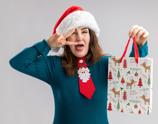 领带快乐的成年白人妇女戴着圣诞帽 打着圣诞领带 伸出舌头 拿着一个纸礼盒 背景是白色的 有复印空间成人舌头背景