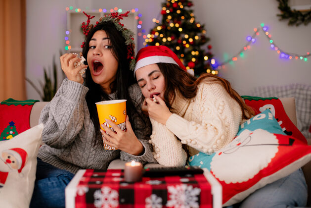 快乐带着冬青花环的饥肠辘辘的年轻女孩吃着爆米花 看着她困倦的朋友坐在扶手椅上 在家里享受圣诞节时光圣诞快乐看享受