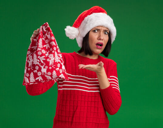 麻袋无知的年轻女孩戴着圣诞帽 拿着圣诞礼物袋 指着隔离在绿色墙上的圣诞礼物袋帽子指着年轻人