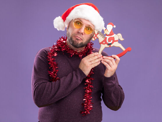 摇摆笨手笨脚的中年男子戴着圣诞老人帽 脖子上戴着金箔花环 戴着眼镜 把圣诞老人抱在摇摇木马上 小雕像在紫色的墙上与它的头相接触花环马头