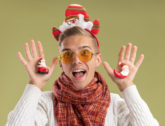 橄榄印象深刻的年轻帅哥戴着圣诞老人的头带和围巾拿着圣诞老人的圣诞饰品看着孤零零的橄榄绿墙上圣诞装饰品戴着