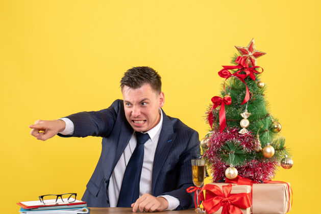 男人正面图是一个年轻人指着坐在桌子旁的圣诞树和黄色墙上的礼物公司坐着的男人