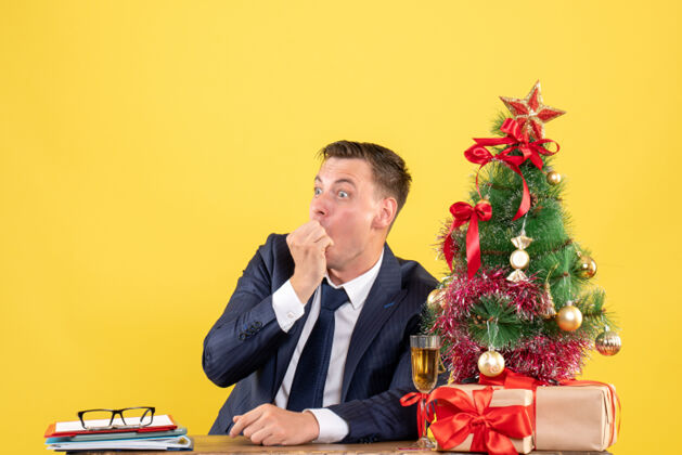 人前视图惊讶的人坐在圣诞树附近的桌子上 并提出了黄色墙壁上的自由空间圣诞节公司办公室