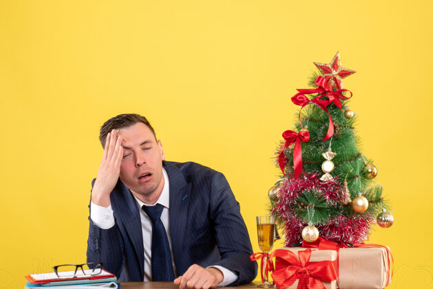 公司失望的男人抱着头坐在圣诞树旁的桌子前 黄色的礼物西装男人黄色